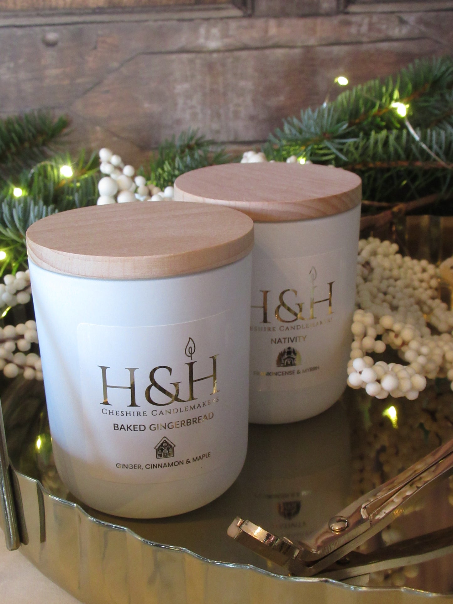 Close up Lifestyle image of Baked Gingerbread candle with Nativity candle by H&H Cheshire Candlemakers in white jars with wooden lids on a gold tray with gold wick trimmers, snowberry branch, fir tree greenery with fairy lights in the background. Label is white square with gold foil text reads Baked Gingerbread Ginger Cinnamon and Maple with a small gingerbread house gold motif.