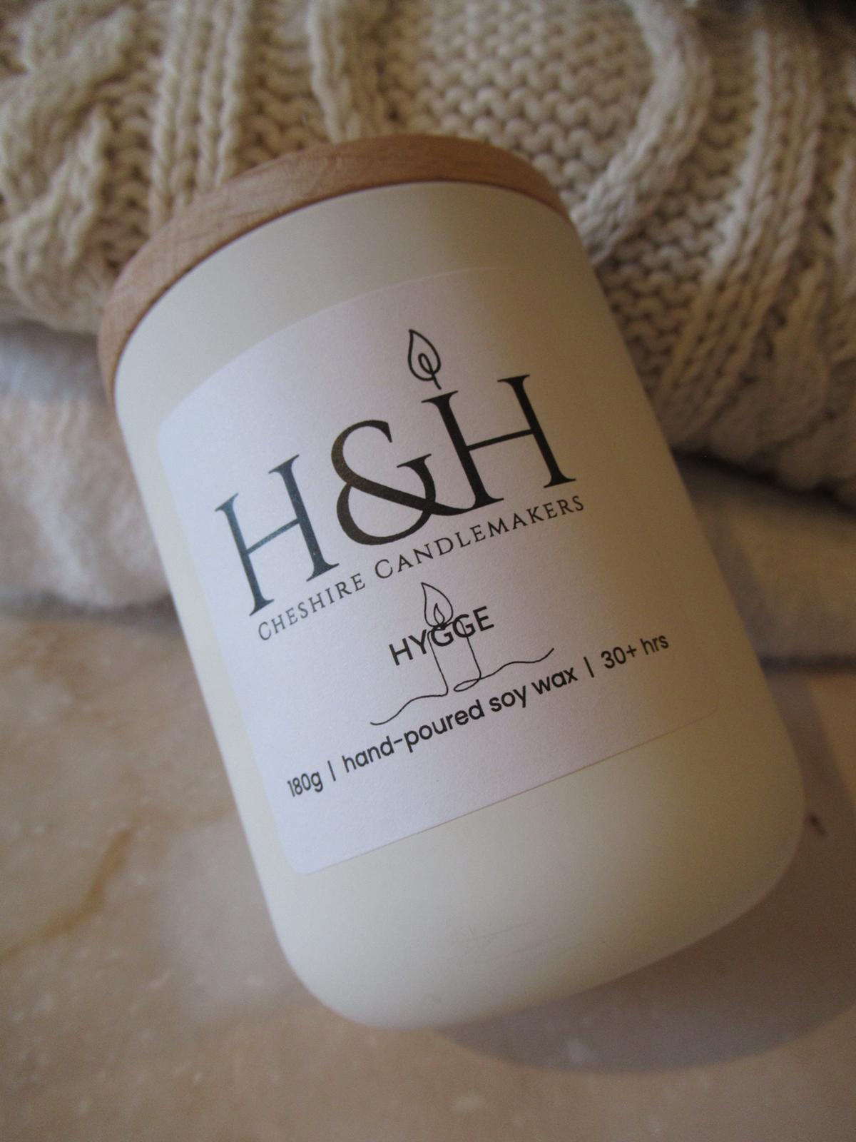 Closeup tilted image of Hygge candle on marble surface in white jar with pale wooden lid. The white label is square with rounded corners and black type incl. H&H Cheshire Candlemakers logo with flame icon and Hygge name with a line drawing candle icon. Text reads 180g | hand-poured soy wax | 30hrs . A cream cable sweater and cashmere cream sweater sit behind candle for cosy neutral autumn vibe display.