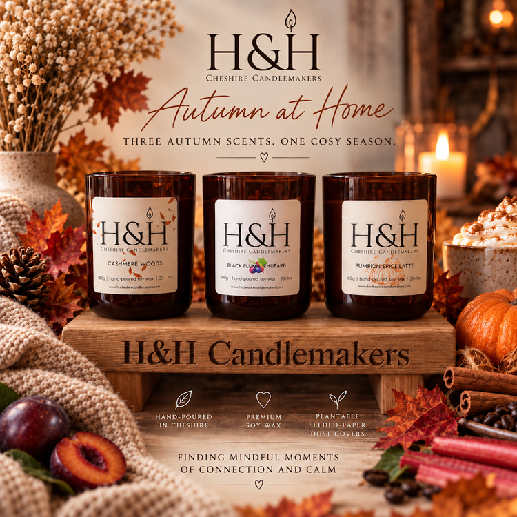 Hero image of 3 autumn H&H candles on a wooden plinthe in a row in amber jars surrounded by blankets, a pumpkin, vase of dried flowers, plums etc