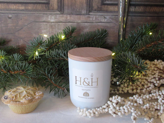 Lifestyle image of Nativity Christmas candle with Nativity candle in a white jar with wooden lid on white velvet with a mince pie, snowberry branch, fir tree greenery with fairy lights in the background. Label is white square with gold foil text reads Nativity Frankincense & Myrrh with a small Nativity scene gold motif