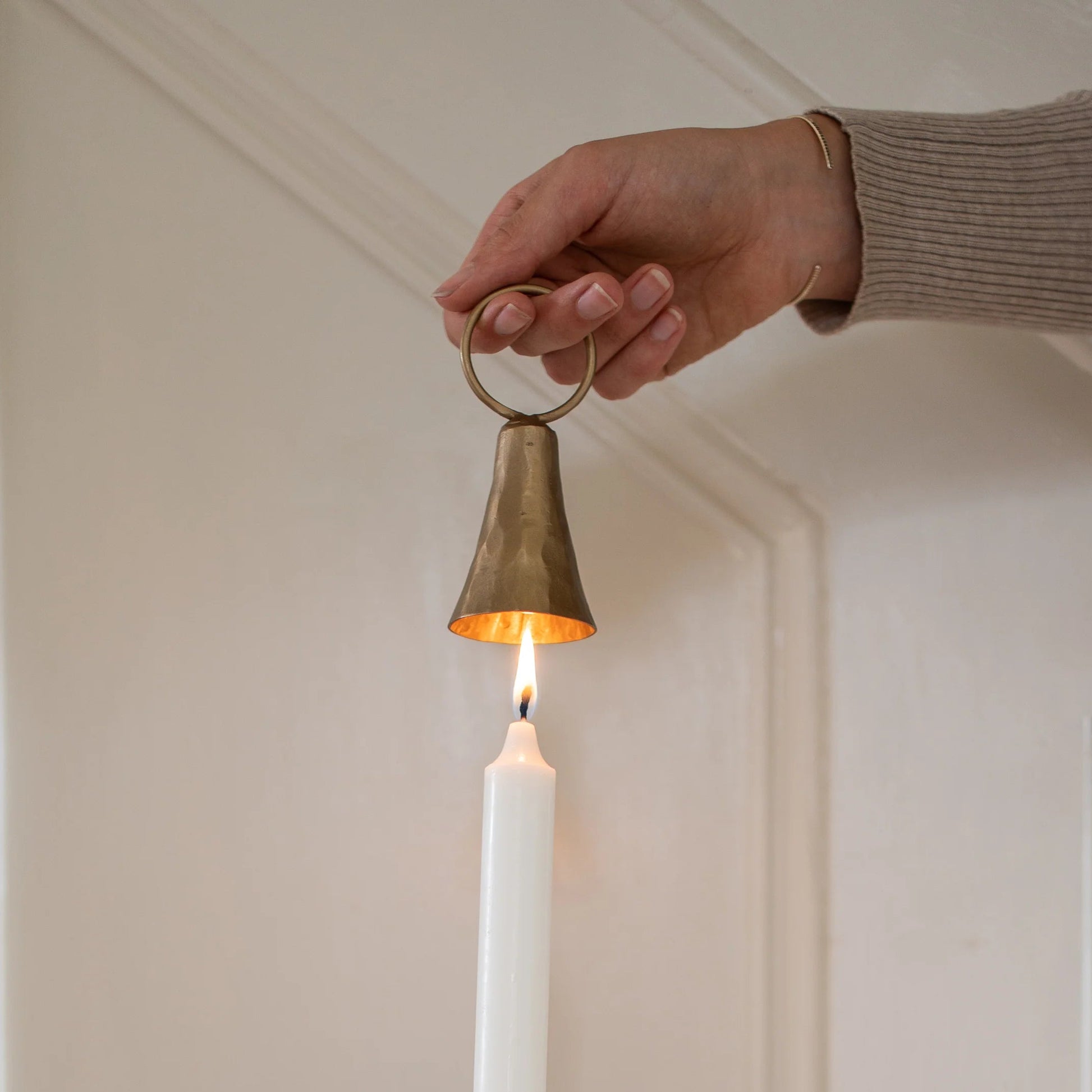Close up of person using brass bell-shaped candle snuffer to extinguish a cream taper candle. Colours are cream background.