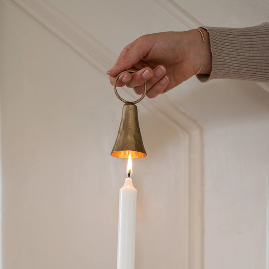 Close up of person using brass bell-shaped candle snuffer to extinguish a cream taper candle. Colours are cream background.