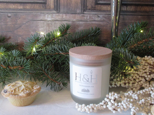 Christmas candle in white frosted jar with pale wooden lid on white velvet tablecloth with fir branch behind and  fairy lights, snowberry branch to the right and a whole mince pie to the left. Background is dark wooden door. Label is white square with silver foil H&H Cheshire Candlemakers logo with flame icon and fir trees icon. Text reads Nordmann Fir Pine, Eucalyptus and Cedar. 