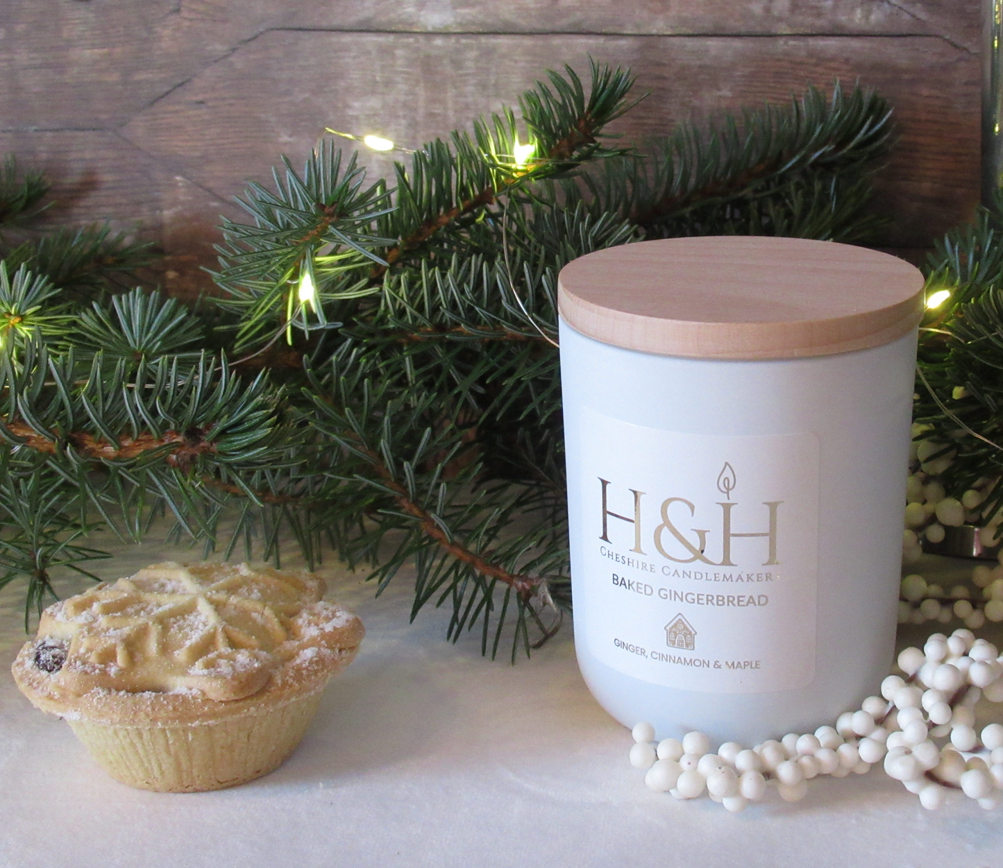 Lifestyle photo of Baked Gingerbread soy wax candle by H&H Cheshire Candlemakers in a white jar with a wooden lid, a white square label with rounded corners and gold foil text. H&H CHeshire Candlemakers logo with flame icon and tiny gingerbread house motif, styled with a mince pie on the left, festive greenery, fairy lights on white velvet cloth.