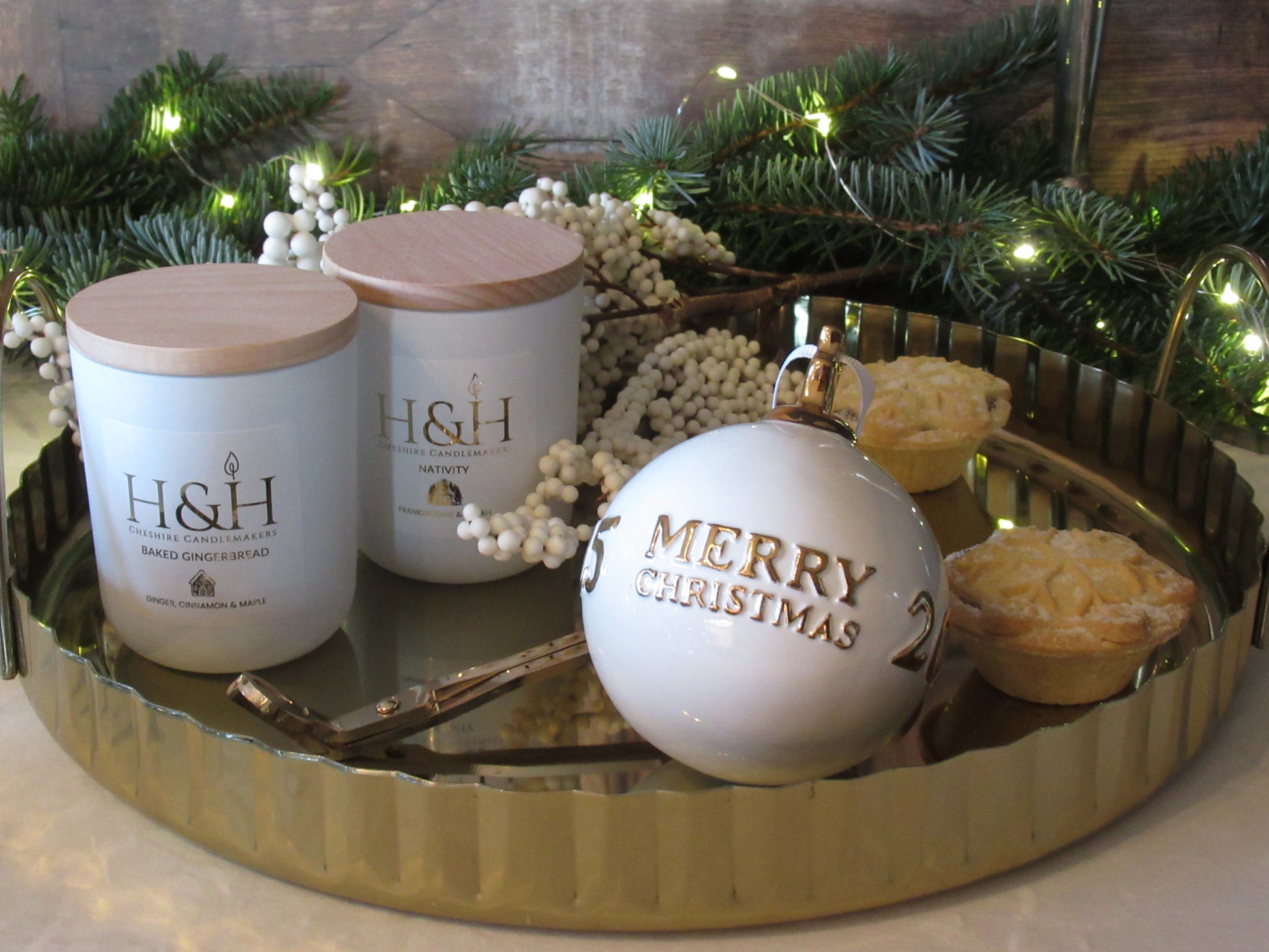 Lifestyle image of Baked Gingerbread candle with Nativity candle by H&H Cheshire Candlemakers in white jars with wooden lids on a gold tray with gold wick trimmers, 2 mince pies, snowberry branch, fir tree greenery with fairy lights in the background and a white and gold Merry Christmas bauble. Label is white square with gold foil text reads Baked Gingerbread Ginger Cinnamon and Maple with a small gingerbread house gold motif.