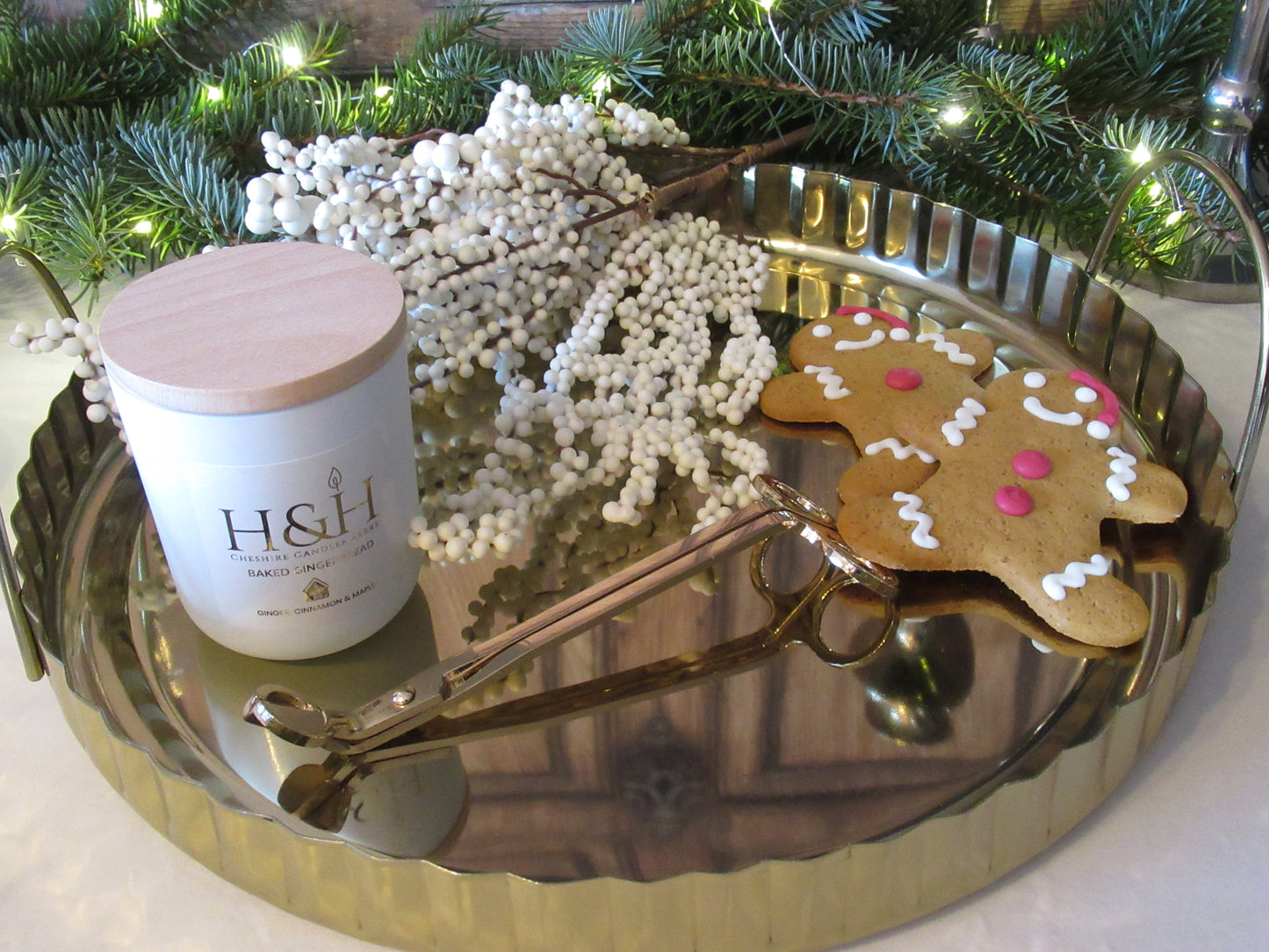 Lifestyle image of Baked Gingerbread candle by H&H Cheshire Candlemakers in a white jar with a wooden lid on a gold tray with gold wick trimmers, 2 gingerbread men, snowberry branch, fir tree greenery with fairy lights in the background. Label is white square with gold foil text reads Baked Gingerbread Ginger Cinnamon and Maple with a small gingerbread house gold motif.