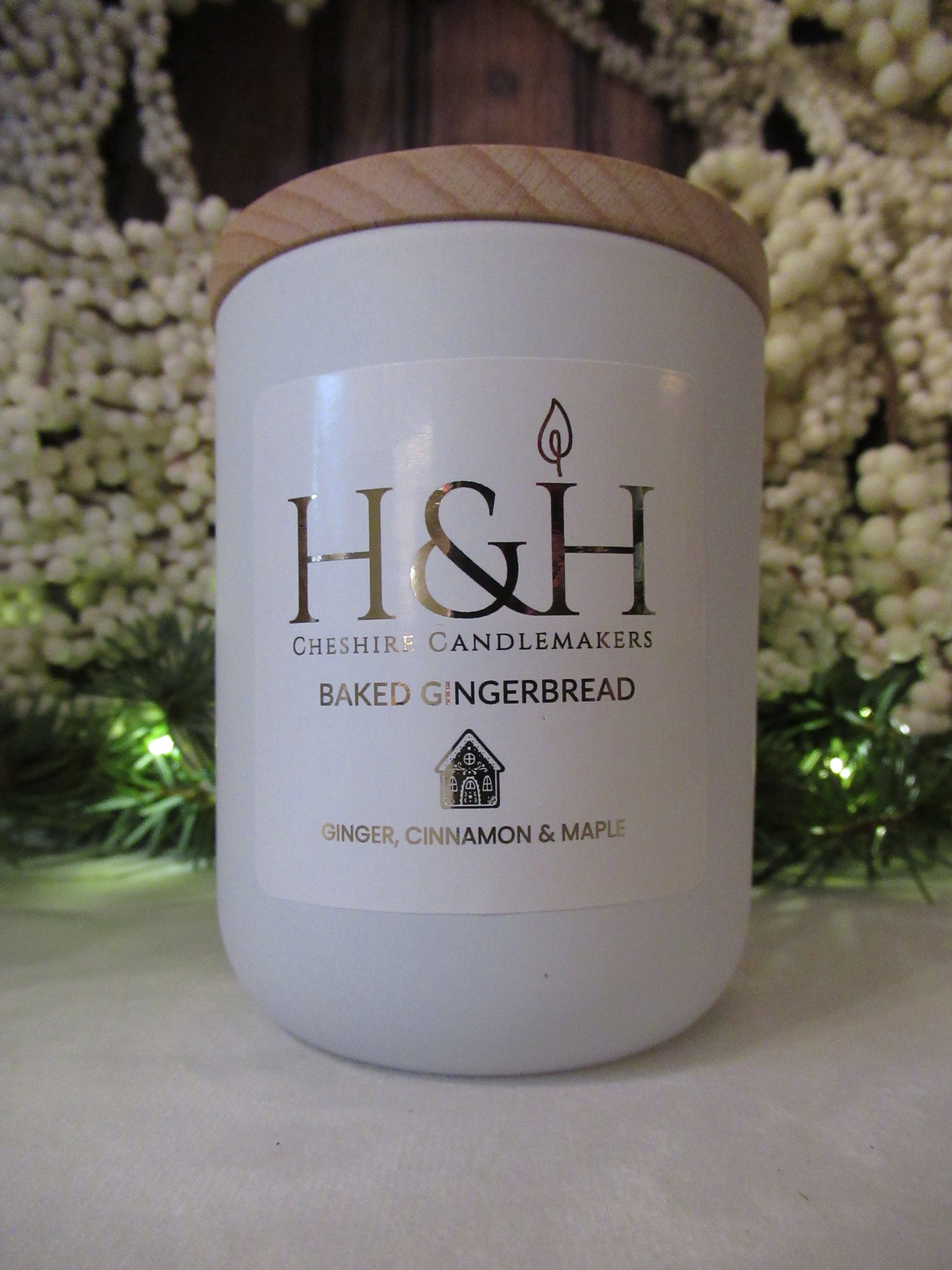 Close up of Baked Gingerbread soy wax candle by H&H Cheshire Candlemakers in a white jar with a wooden lid, a white square label with rounded corners and gold foil text. H&H CHeshire Candlemakers logo with flame icon and tiny gingerbread house motif, styled with festive greenery and soft lights on white velvet cloth.