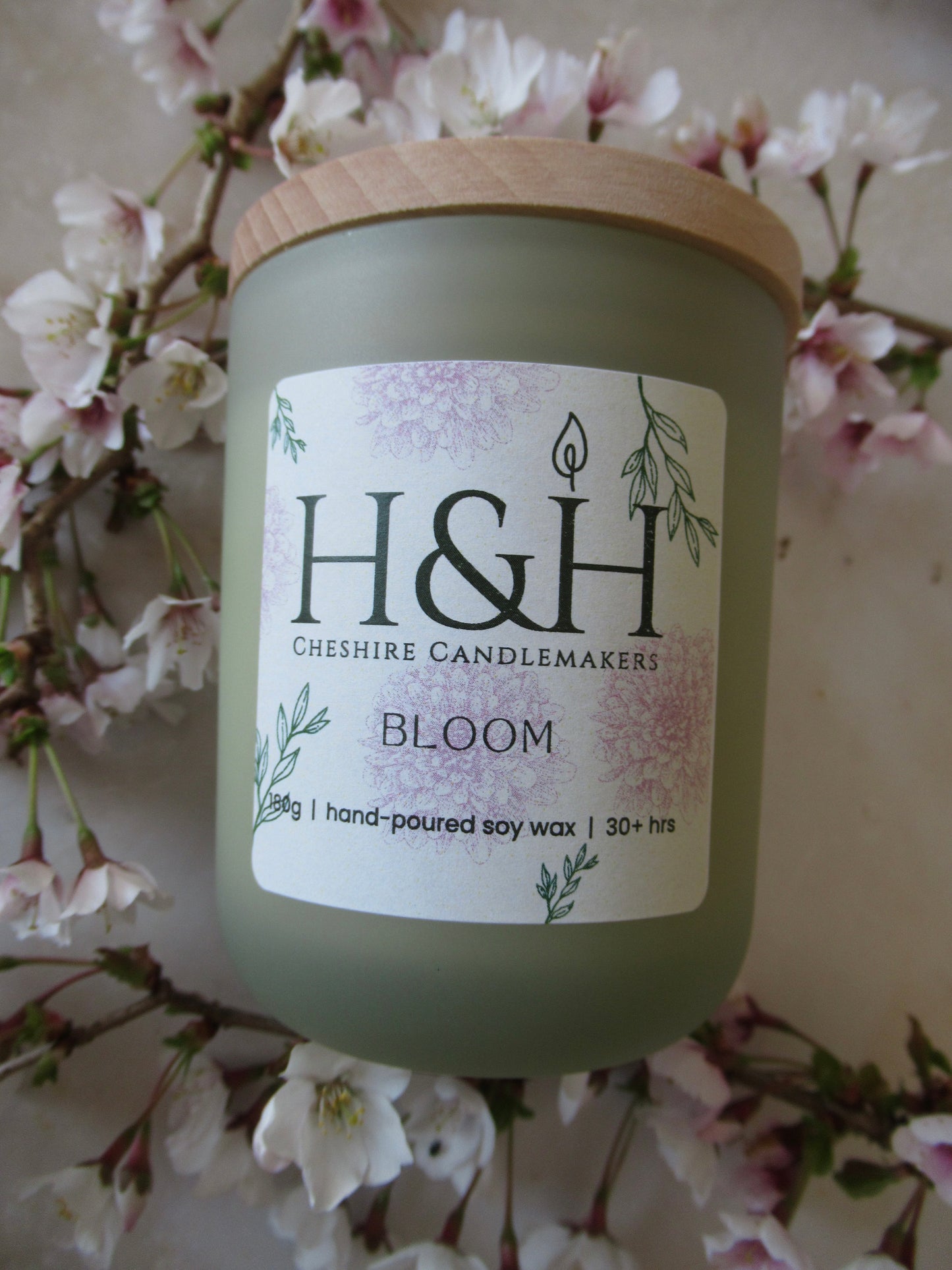 Close up flatlay photo of Bloom candle slightly tilted to right in sage green jar with pale wooden lid surrounded by small blossoms flowers on a marble surface. The label is cream square with rounded corners with pale pink flowers and green leaves motif and the dark green H&H Cheshire Candlemakers logo. Text reads Bloom 180g|handpoured soy wax|30hrs +