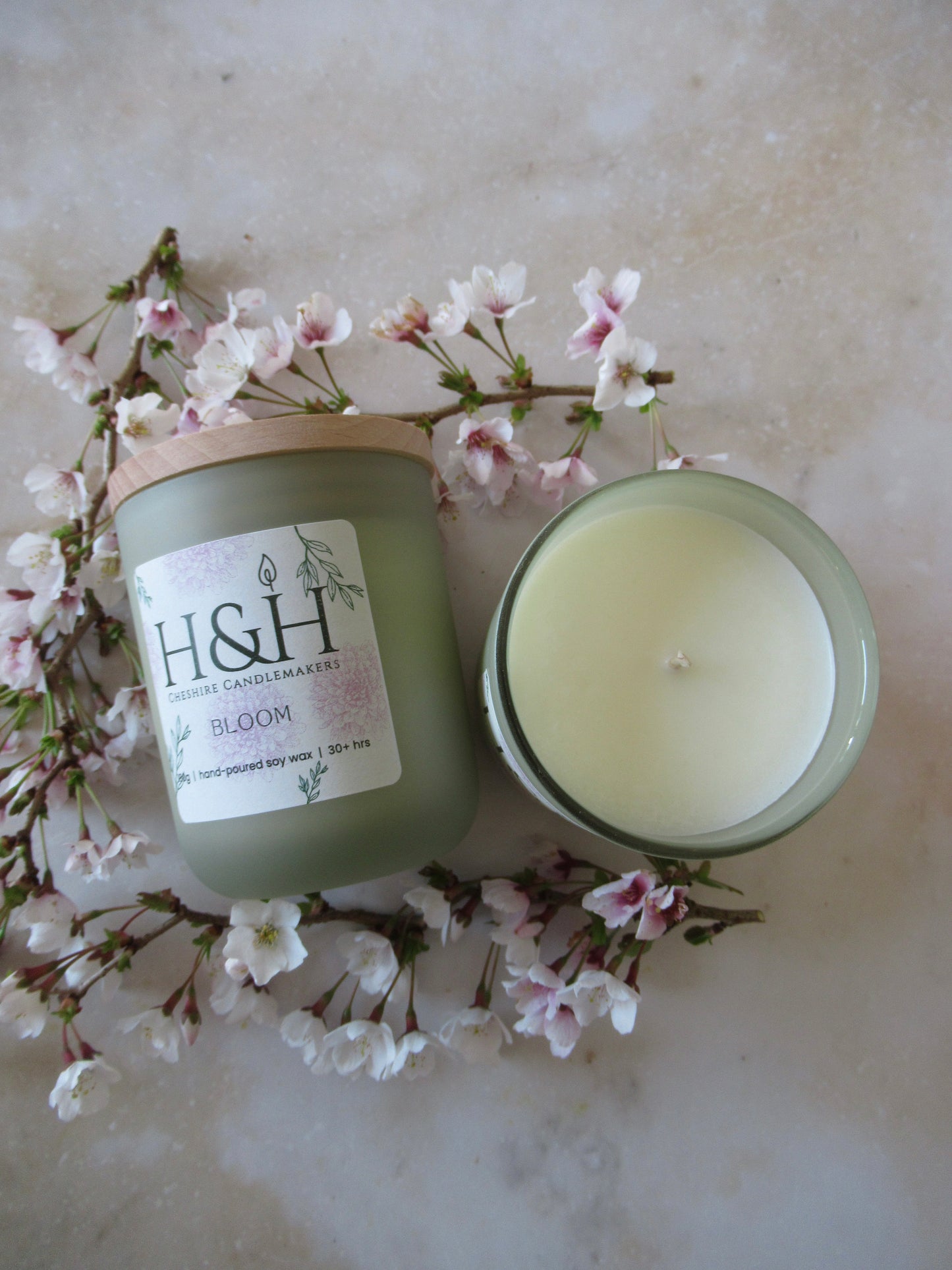 Close up flatlay photo of 2 Bloom candles in sage green jar with pale wooden lids surrounded by small blossoms flowers on a marble surface. One candle has no lid showing the wax suface and wick from above. The label is cream square with rounded corners with pale pink flowers and green leaves motif and the dark green H&H Cheshire Candlemakers logo. Text reads Bloom 180g|handpoured soy wax|30hrs +