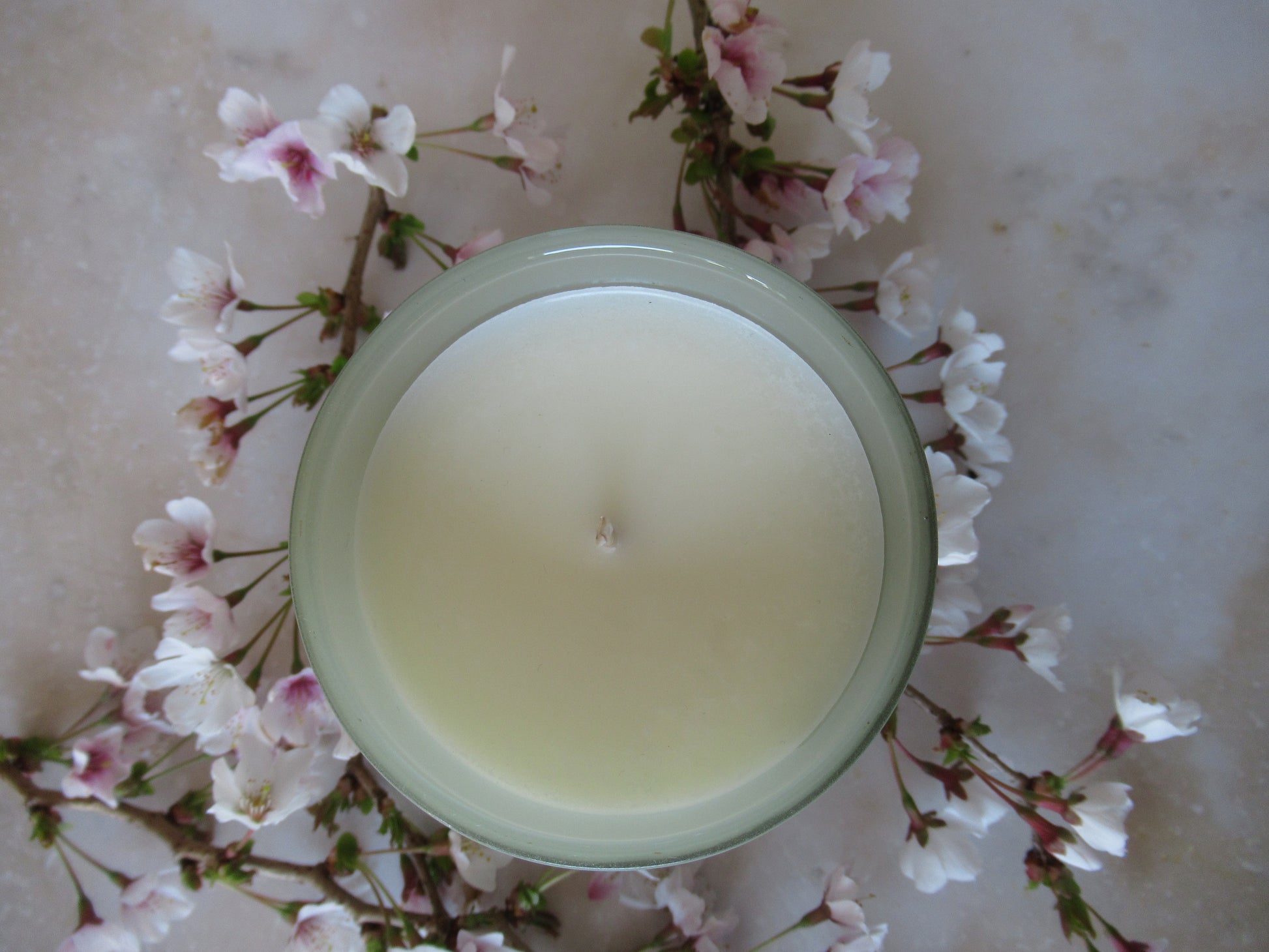 Close up flatlay photo of Bloom candle from above in sage green jar surrounded by small blossoms flowers on a marble surface. The candle has no lid showing the wax suface and wick from above. 