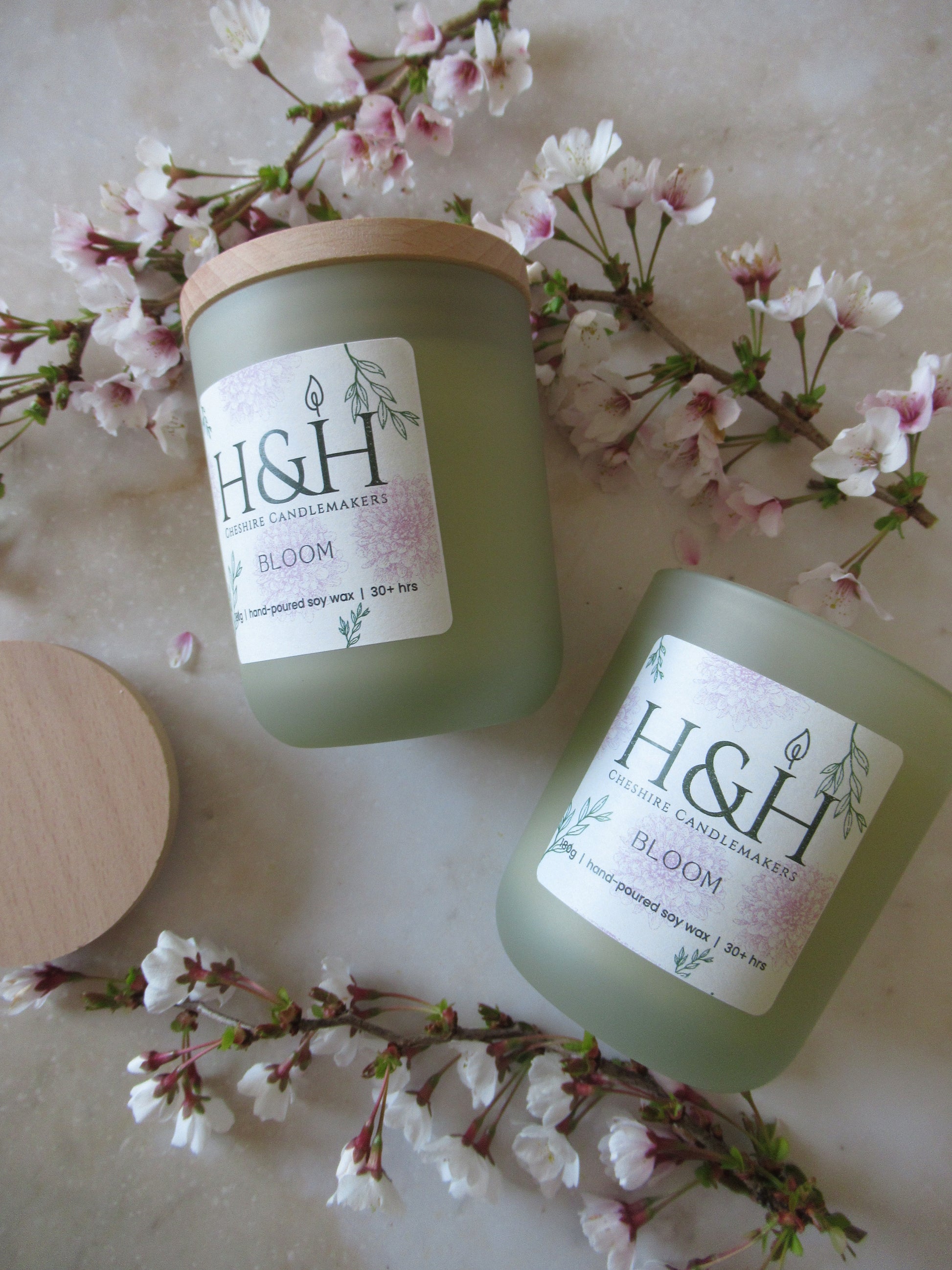 Close up flatlay photo of 2 Bloom candles in sage green jar with pale wooden lids surrounded by small blossoms flowers on a marble surface. The label is cream square with rounded corners with pale pink flowers and green leaves motif and the dark green H&H Cheshire Candlemakers logo. Text reads Bloom 180g|handpoured soy wax|30hrs +