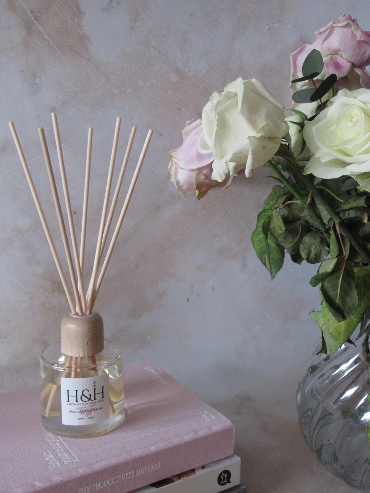Lifestyle photo of Wild Bramble & Rose vegan reed diffuser in a clear glass bottle with natural wooden reeds. Displayed on pink books next to a vase of white and pink roses on a marble surface.