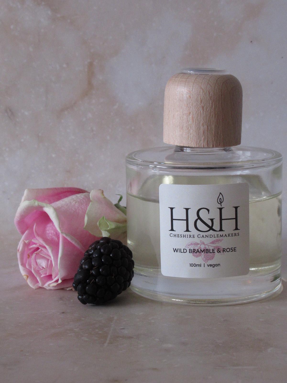 Close-up of Wild Bramble & Rose vegan reed diffuser by H&H Cheshire Candlemakers, in a clear glass bottle with a natural wooden cap, styled with a pink rose and fresh blackberry on a marble surface.