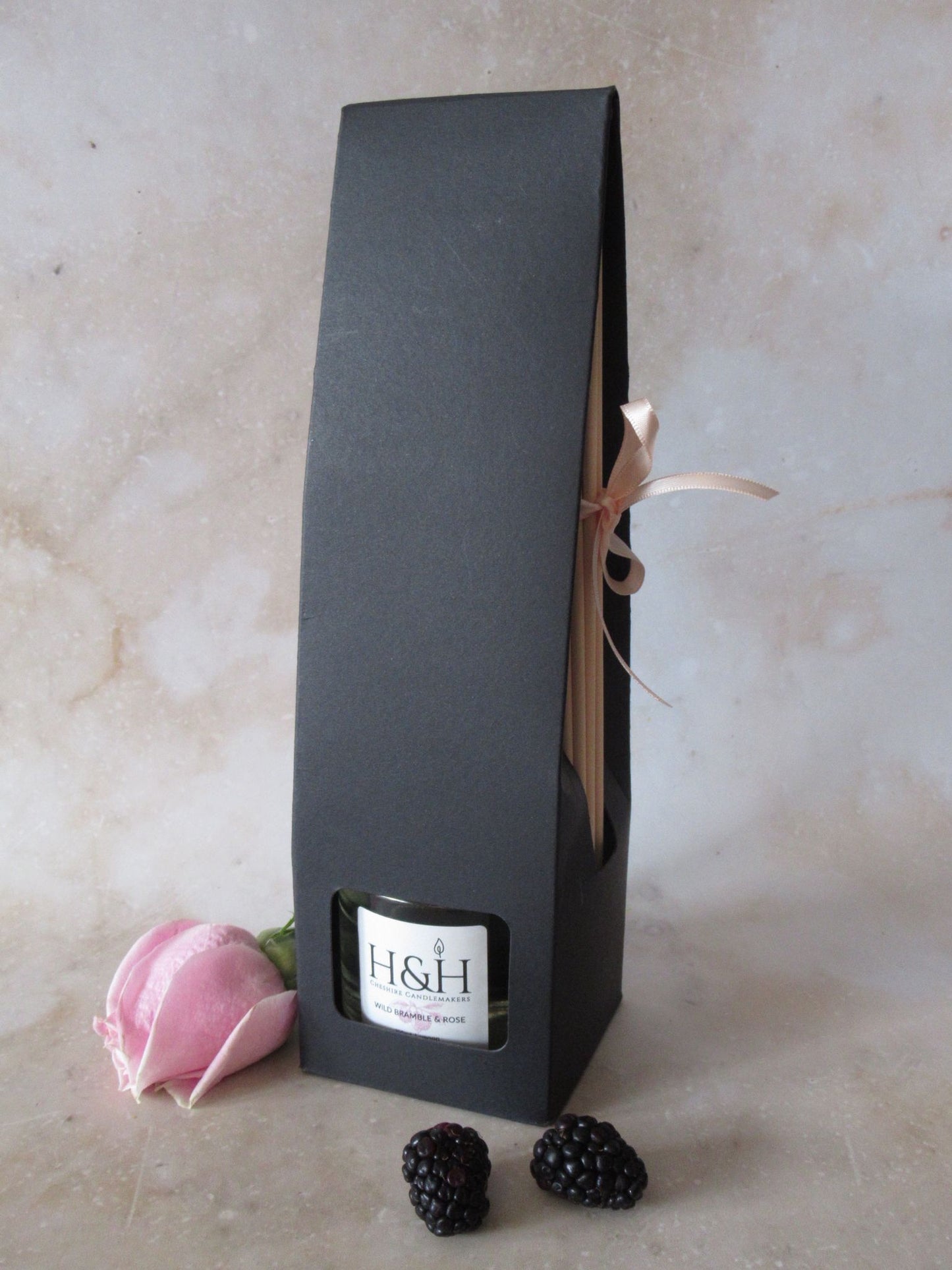 Wild Bramble & Rose vegan reed diffuser in black box packaging with window for bottle and a soft pink ribbon tying reeds on the side. Styled with a pink rose and fresh blackberries on a marble background.