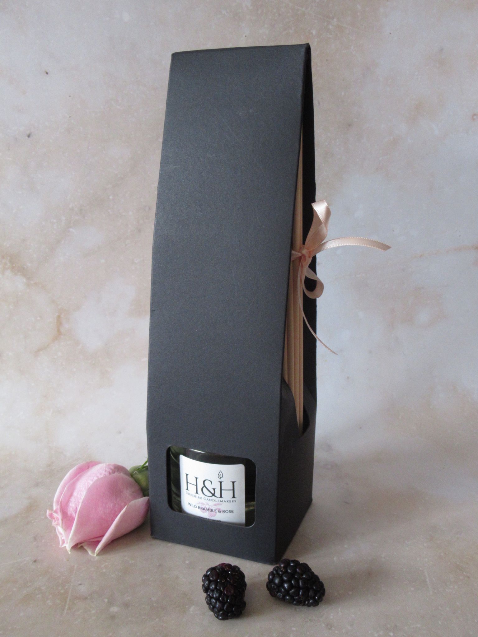 Wild Bramble & Rose vegan reed diffuser in black box packaging with window for bottle and a soft pink ribbon tying reeds on the side. Styled with a pink rose and fresh blackberries on a marble background.