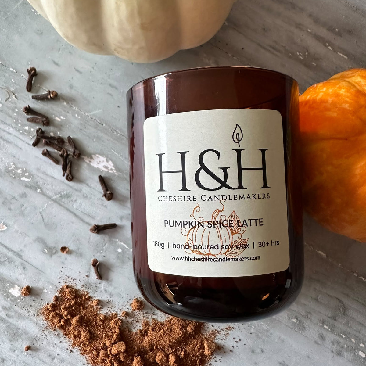 An amber glass candle jar labeled H&H Cheshire Candlemakers Pumpkin Spice Latte, styled on a gray surface with a white pumpkin, orange pumpkin, cloves, and cinnamon powder.