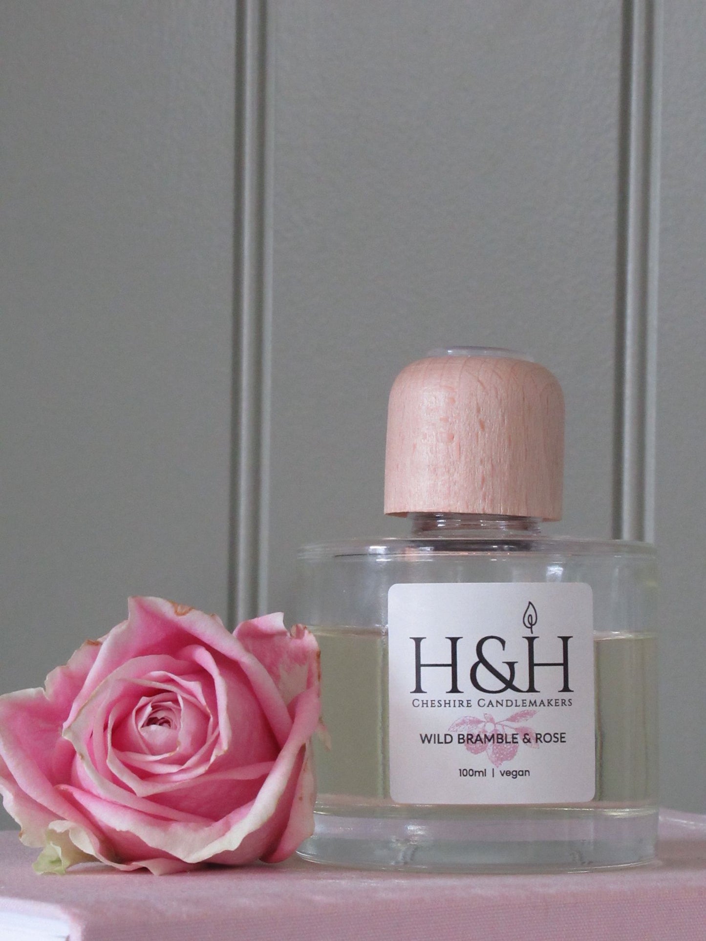 Close-up of Wild Bramble & Rose vegan reed diffuser in a clear glass bottle with a natural wooden cap. Styled with a soft pink rose against a sage green panel backdrop.