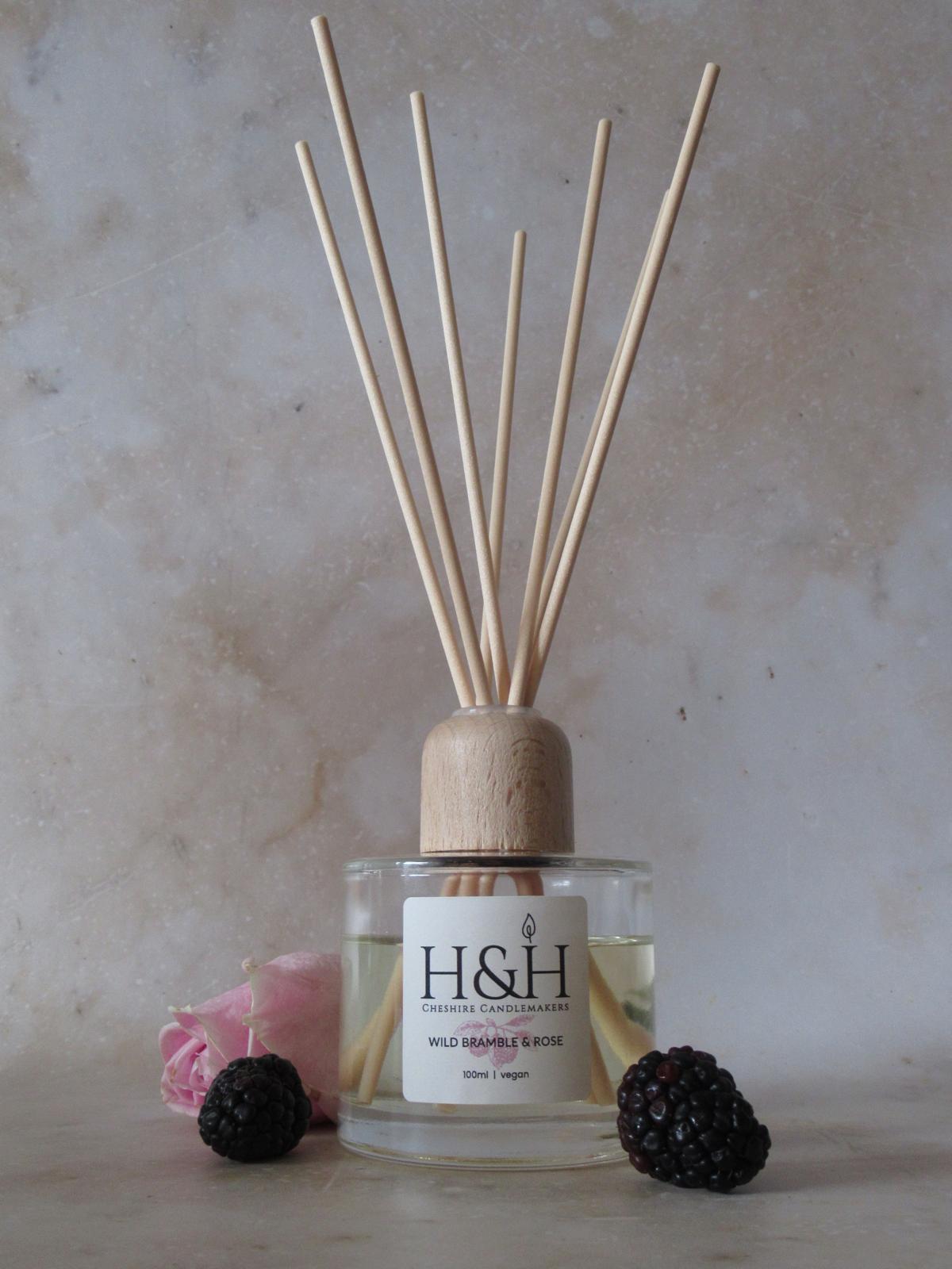 Wild Bramble & Rose reed diffuser with clear glass bottle, natural wooden cap, and reeds. Styled with 2 blackberries and a pink rose on a marble background.