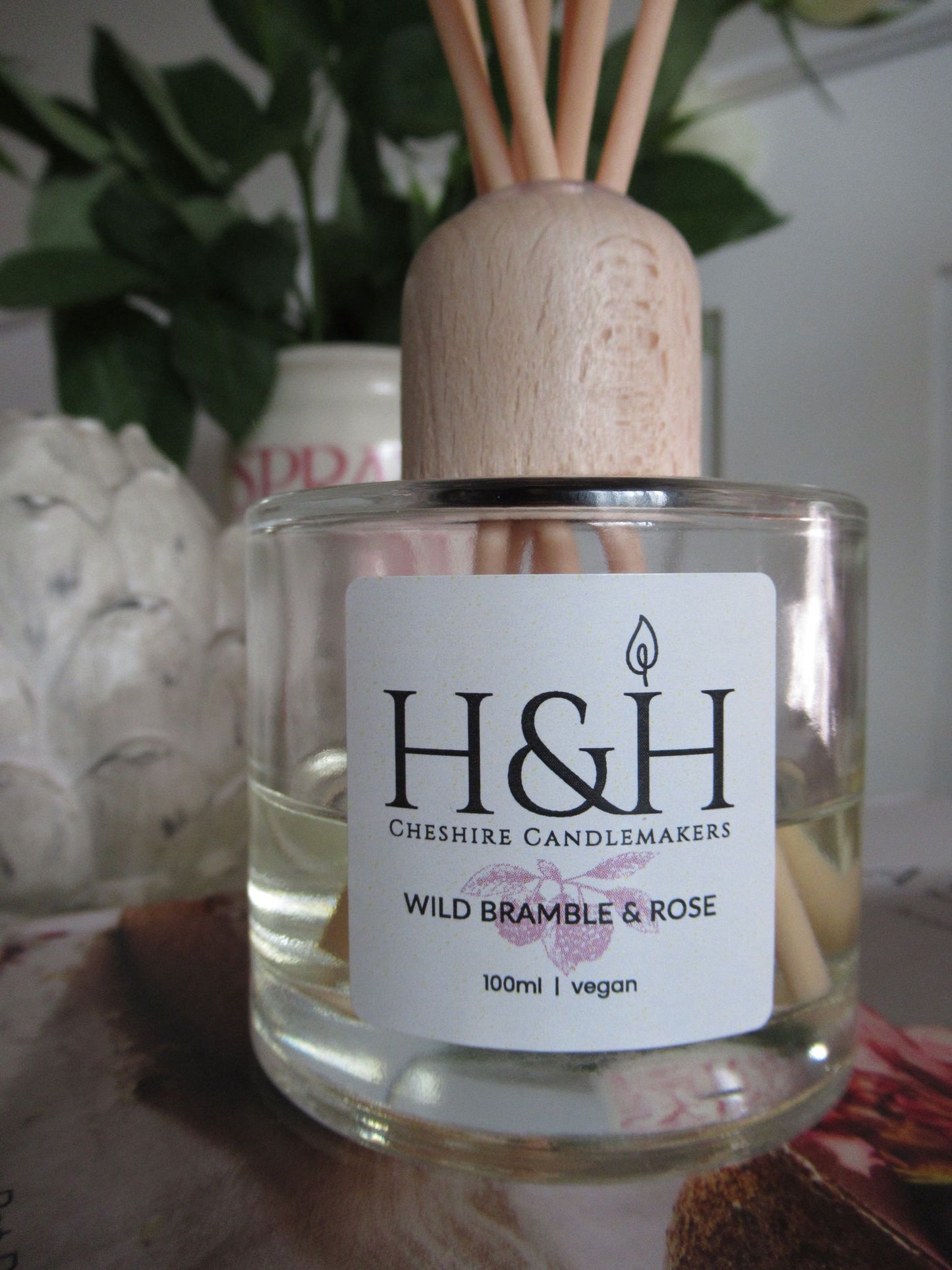 Close up lifestyle photo of glass bottle and label of Wild Bramble & Rose diffuser with 'H&H Cheshire Candlemakers Wild Bramble & Rose and 100ml | Vegan' text on label.