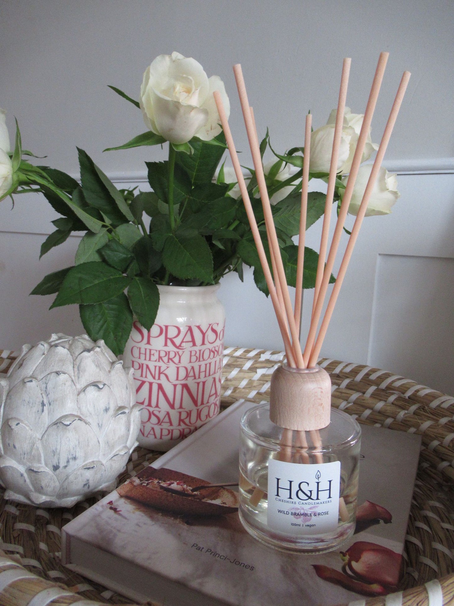 Lifetyle image of Wild Bramble & Rose diffuser on a wicker tray with cream roses in a ceramic vase and a wooden artichoke ornament. 