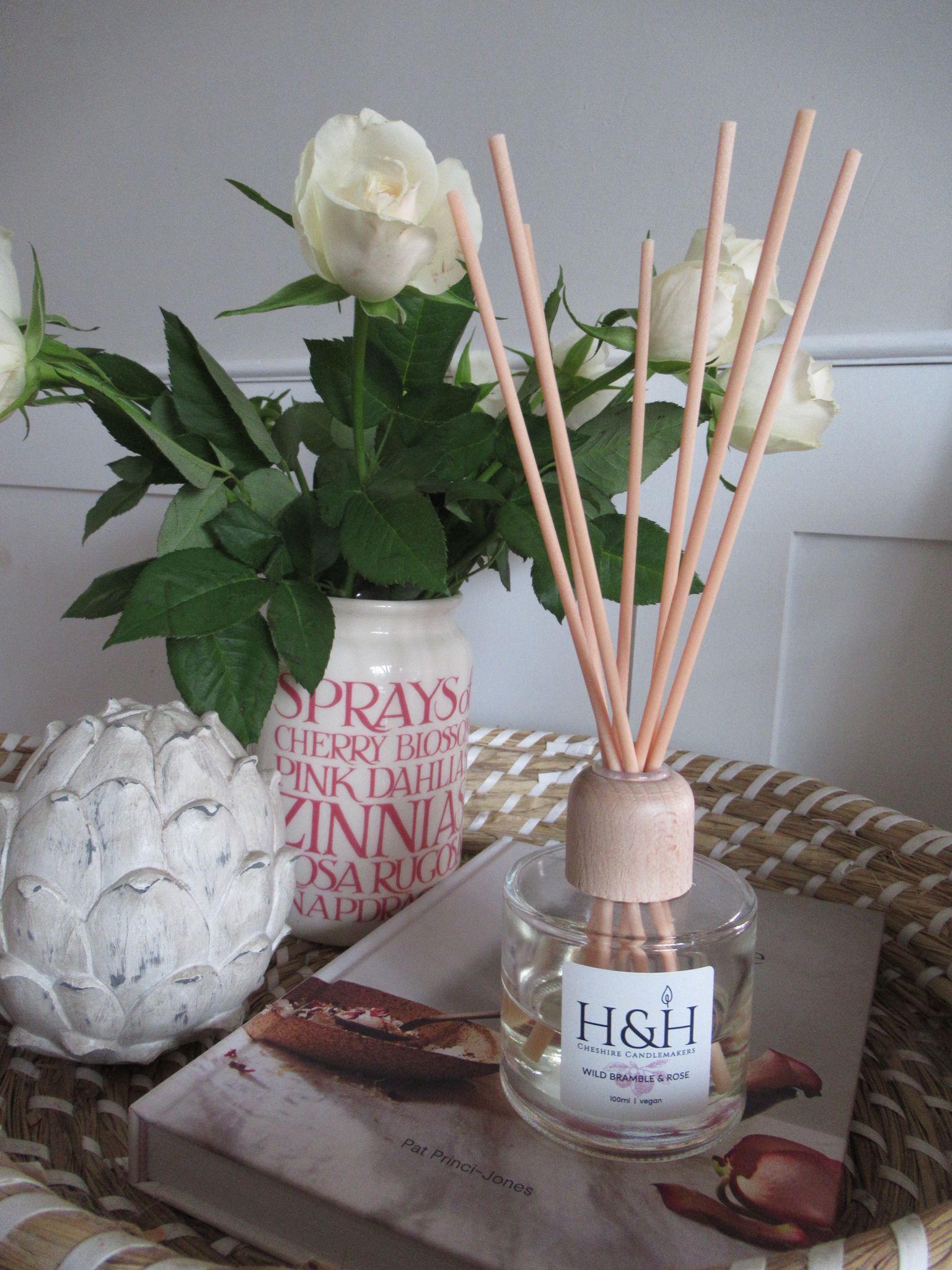 Lifetyle image of Wild Bramble & Rose diffuser on a wicker tray with cream roses in a ceramic vase and a wooden artichoke ornament. 