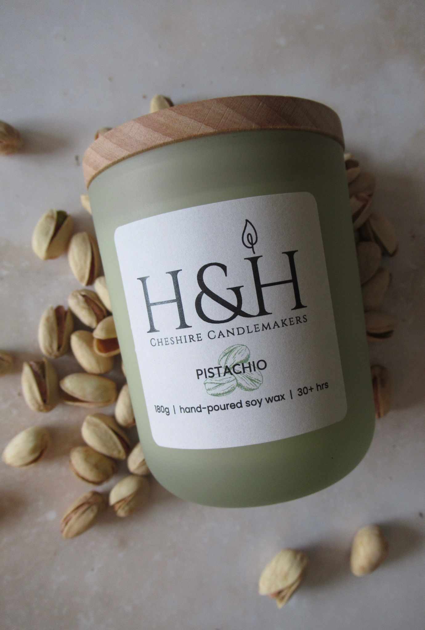 Flatlay of Pistachio soy wax candle in sage green jar with wooden lid, surrounded by pistachio nuts on marble. Label reads: 180g | hand-poured soy wax | 30hrs+.