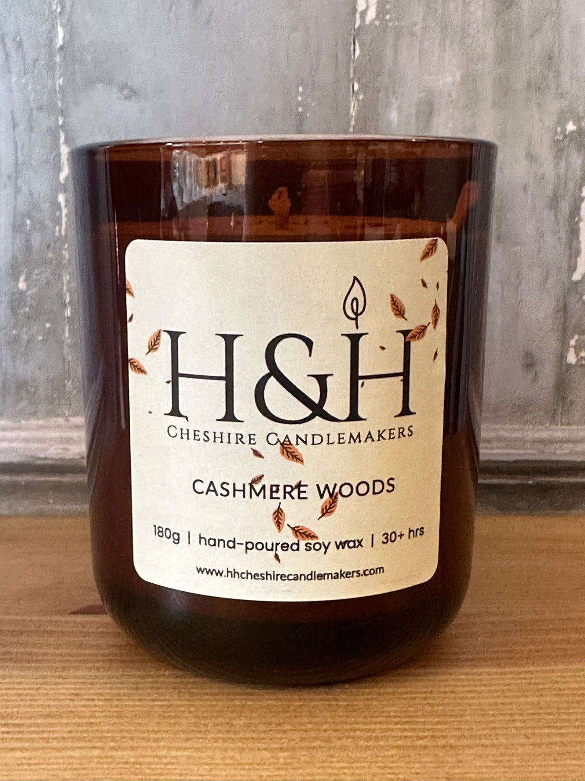 An amber glass jar candle with a cream label reading 'H&H Cheshire Candlemakers Cashmere Woods' on a wooden surface against a gray textured wall.