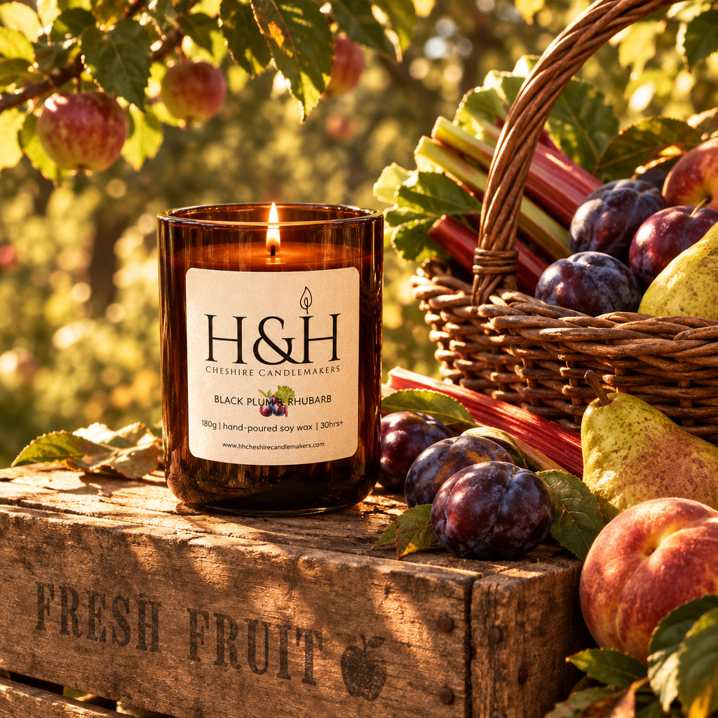 AI Image of an amber glass jar candle with a white label reading H&H Cheshire Candlemakers and Black Plum & Rhubarb, placed a wooden fruit crate in a sunlit orchard next to a basket of plums, pears, peaches and rhubarb.