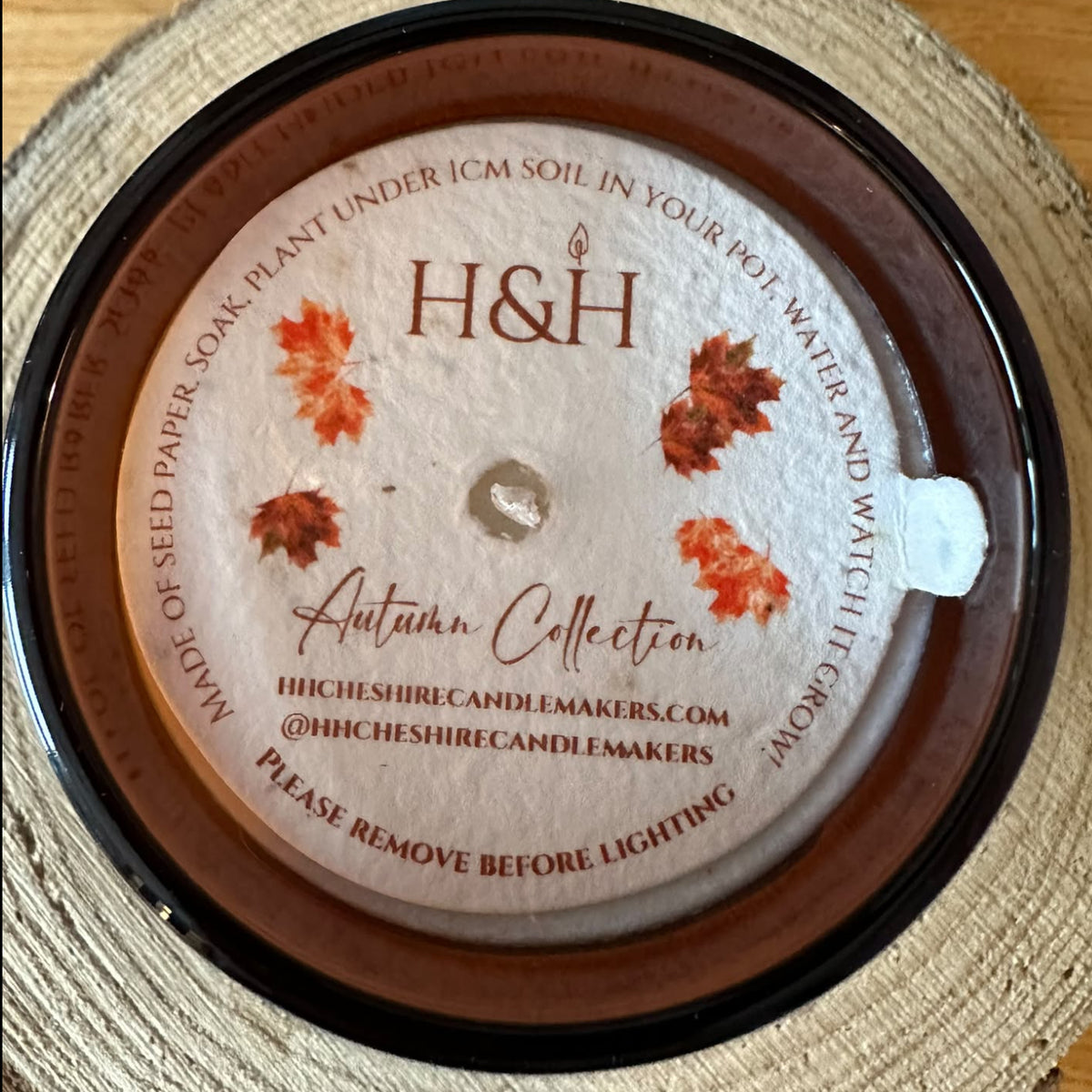 A top-down view of an H&H Autumn Collection soy wax candle in a round amber jar with round seeded paper dust cover on top, resting on a circular wood slice coaster over a wooden tabletop.