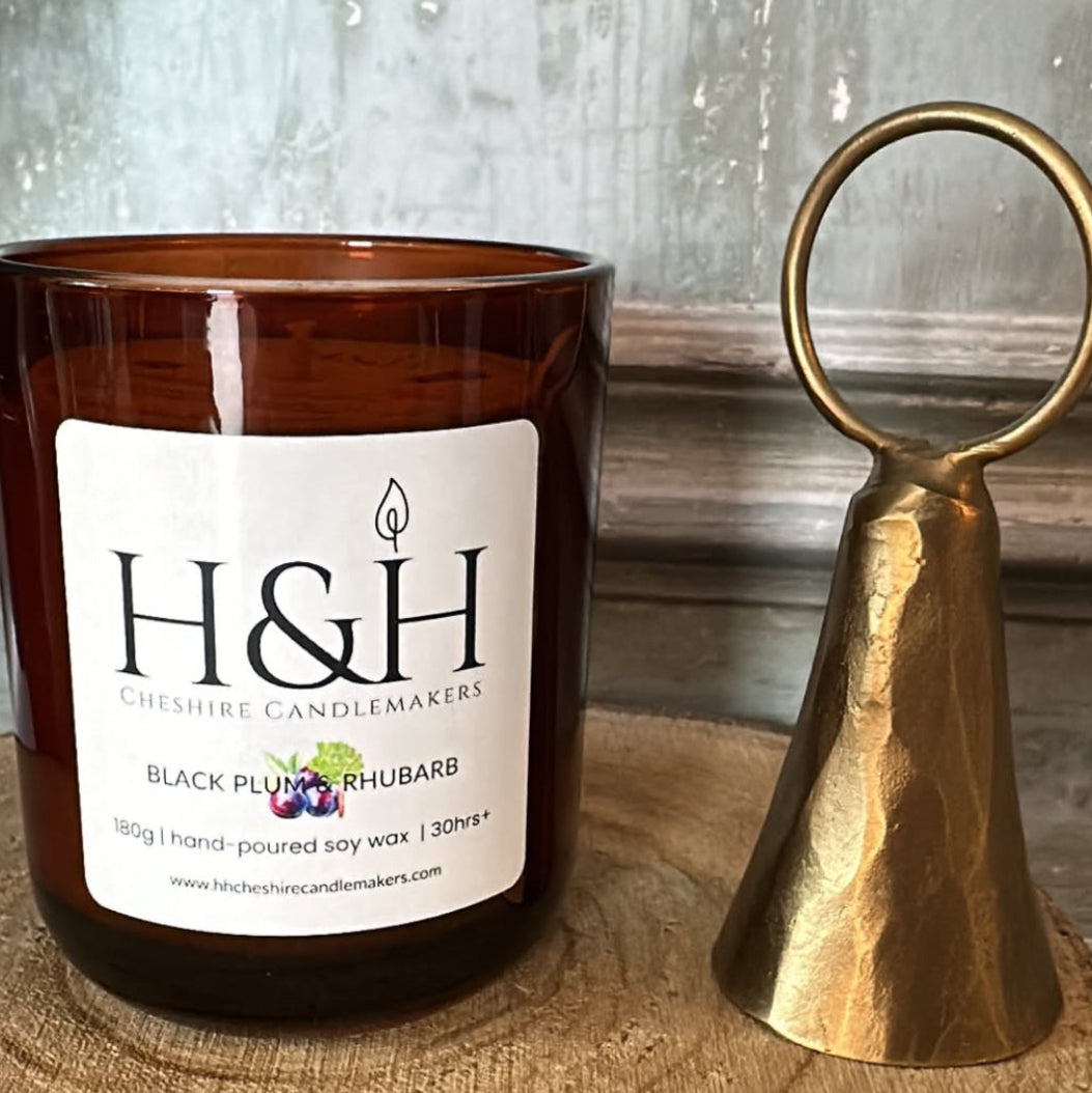An amber glass candle jar with a white H&H Candlemakers label sits on a rustic wood slice next to a small hammered brass bell snuffer against a weathered gray wooden backdrop.