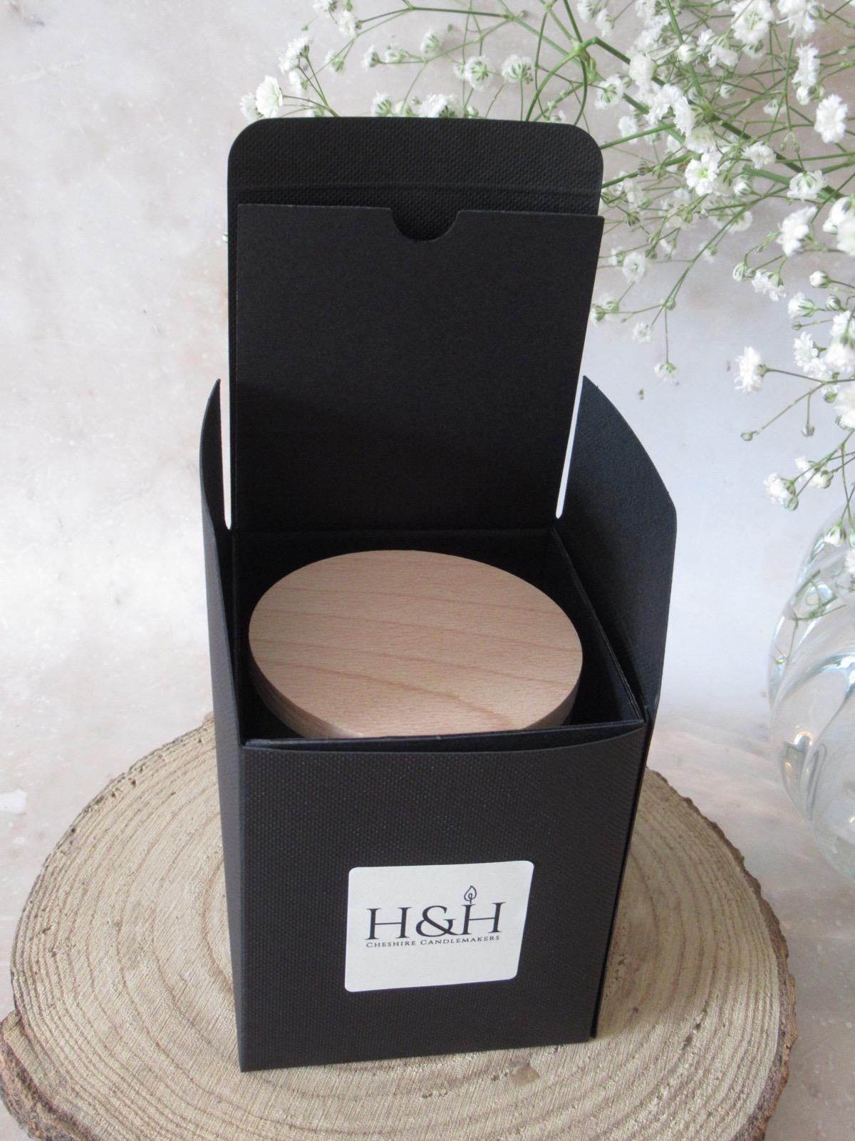 Open black gift box with a candle inside showing the wooden lid on a wooden slice surface with white gypsophila in the background