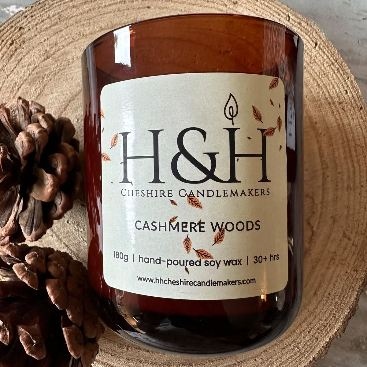 An amber glass jar candle with a cream label reading H&H Cheshire Candlemakers Cashmere Woods, styled on a round woven coaster with pinecones beside it.