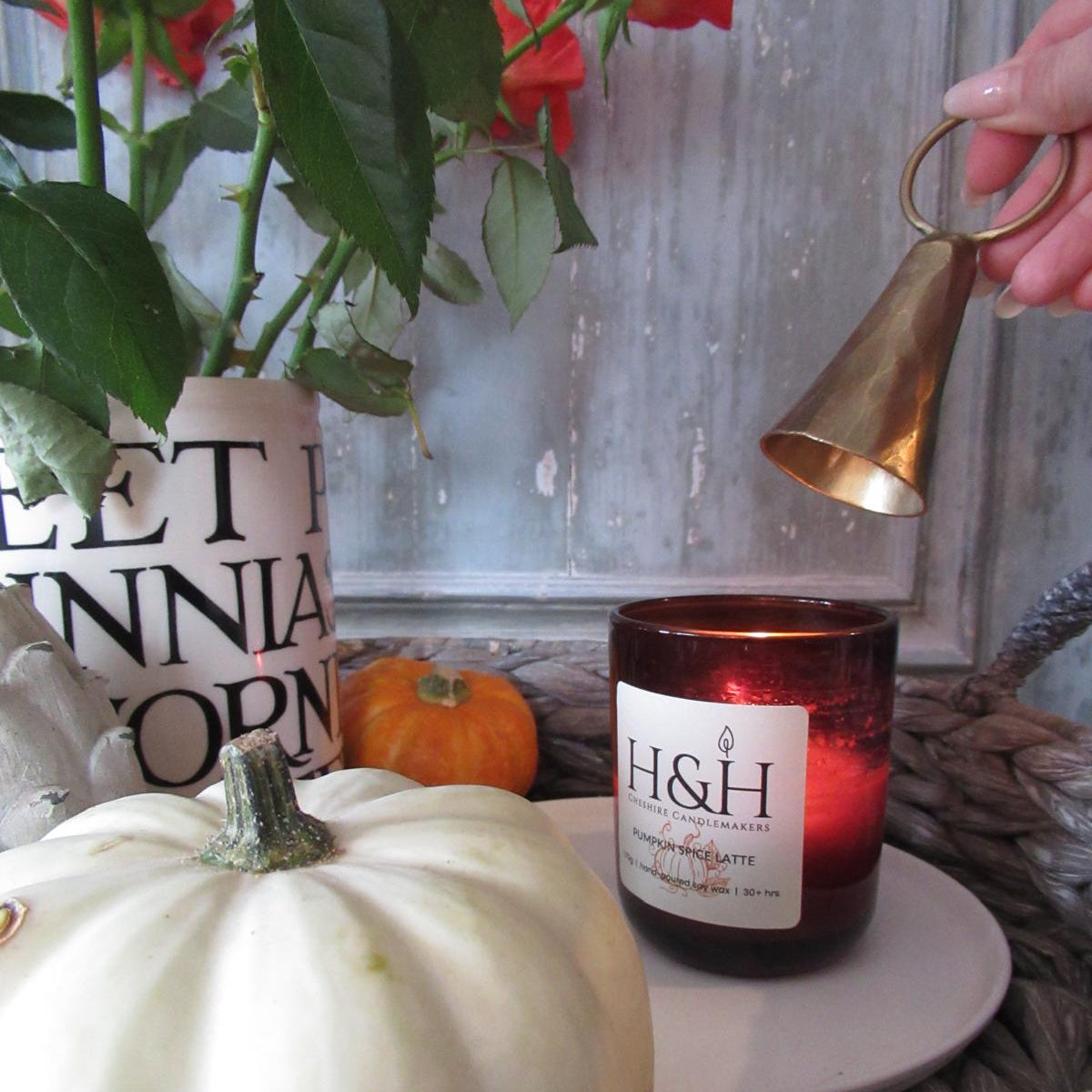 A styled autumn still life featuring a white pumpkin in the foreground, an amber candle in a glass jar on a white plate, a cream ceramic jar with orange roses, and a white wooden artechoke in a woven basket. Someone is holding a brass bell-shaped candle snuffer over the candle.