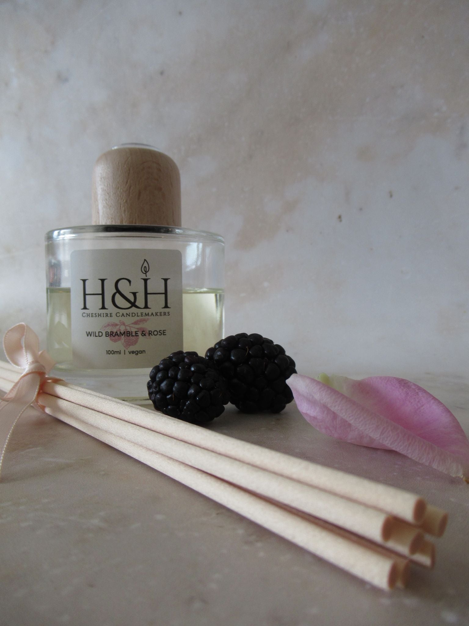 Clear round glass bottle of Wild Bramble & Rose vegan reed diffuser with a natural wooden cap, blackberries, pink rose petal, and natural reeds on a marble surface.